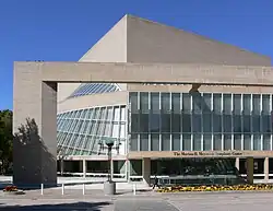 A beige cube rises at an angle around a half-cone made of glass and steel. In front, a square archway overlooks a stone courtyard.