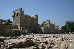 A stone wall intersected by three square towers with the ruins of another building in the foreground