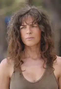 Head and shoulders of an attractive middle aged woman with long curly brown hair, wearing a brown tank top, with a sad frown on her face.