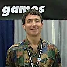 A Caucasian man with short brown hair and a convention pass on a lanyard around his neck smiles for a camera. Part of the Telltale Games logo is visible in the background.