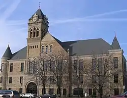 A large three-story stone building built in 1895. Three stories of windows line the front of the building with the two front corners containing cone-shaped roofs that stick out from the main roof. Above the entrance is a large clock tower that is taller than the rest of the building.