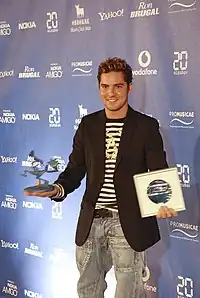 A young blonde man wearing a black-and-white stripped blouse, blazer and jeans is standing in front of a blue background. He has an award in his left hand and a certificate in his right