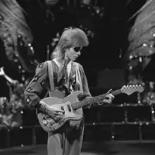 A black and white photo Bowie with longer hair and an eyepatch, holding a guitar and looking down at it