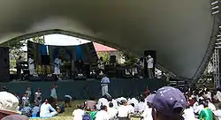 Performers on an outdoor stage with shell-shaped overhang before an audience seated in the sun.