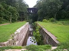 A narrow stone-lined canal lock filled with water surrounded by grass. In the background a path follows the canal at left and a modern bridge crosses it.