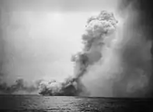 A black and white photograph showing a large cloud of smoke near the sea surface from which issues a towering mushroom cloud angled toward the right side of the photo