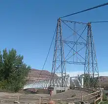 A view of a cable suspension bridge with one metal tower and a wooden deck. The second tower is partially obscured by a cottonwood tree.