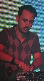 Dimitri from Paris performing on a stage with a multi-coloured background, using a machine and wearing a diamond checked shirt