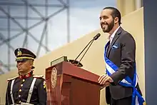 Nayib Bukele standing at a podium, wearing the Salvadoran presidential sash, and speaking to a crowd