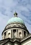 The dome of a building with a green patina, against a blue sky with clouds in it.