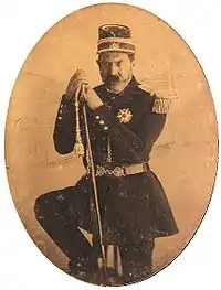 Three-quarters length tintype portrait showing a mustachioed man in military dress uniform and cap with one foot resting on a rock and holding a sheathed sword.