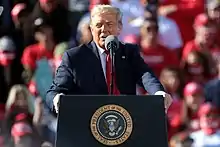 Donald Trump speaking at a podium. Behind him is a crowd.