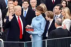 With right hand raised, Donald Trump looks at Chief Justice John Roberts with his back to the camera, as Melania Trump and others watch.