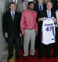 Florida Gators men's basketball coach Billy Donovan, 42 year-old white man shown in navy blue blazer and tie, and his 2007 NCAA championship team, with former Florida Gator Walter Hodge and U.S. President George W. Bush holding Florida Gators jersey "43," at the White House.