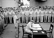 MacArthur is seated a small desk, writing. Two men in uniform stand behind him. A large crowd of men in uniform look on.