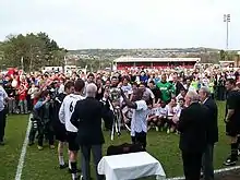 Two men in white football shirts and black shorts receive a silver trophy from an elderly man in a dark blazer. More players look on, as do several more men in blazers and a crowd of spectators of all ages. Several photographers are taking pictures of the presentation.