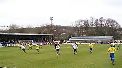 Two teams competing in a football match, one wearing white shirts and black shorts and the other yellow shirts and blue shorts. A stand full of spectators and another low brick building are visible outside the playing area.