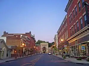 A view of an urban street at dusk with brick buildings on either side and a brown building with a large arched window in the distance at dusk, with the street lamps lit