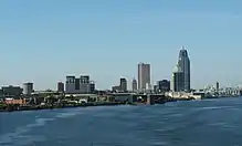 A large city seen from the water on a clear day. The shoreline is fringed with docks above which rise city buildings including several skyscrapers.