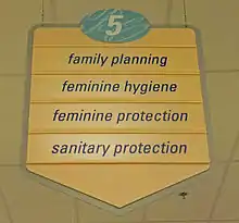 A yellow sign with a pointed bottom. At the top is the number 5 in an oval with a blue background. Below it are the words "family planning", "feminine hygiene", "feminine protection" and "sanitary protection"