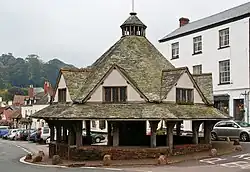 Street scene with houses and shops on the left and an octagonal structure has a central stone pier which supports a heavy timber framework which carries a slate roof with central wooden lantern surmounted by a weather vane. In the distance is a castle.