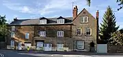 A row of English stone houses with white windows and doors beside a road. There is a stone wall and wooden gate on one side, and several different types of trees around them.