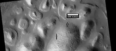 Layers exposed at the base of a group of buttes in Mangala Valles in Memnonia quadrangle, as seen by HiRISE under HiWish program.  Arrows point to boulders sitting in pits.  The pits may have formed by winds, heat from the boulders melting ground ice, or some other process.