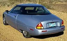 The rear three-quarters view of an electric automobile with a hidden rear wheel and a shown front wheel. Photo is taken in what looks like a desert