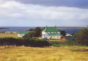 A large, white, two-storey, wooden farm house surrounded by five smaller, rectangular structures and a wooden gazebo. There is a red brick house in the foreground, and another couple of buildings in the background. Beyond is a bay.