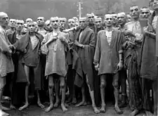 Just-liberated Ebensee concentration camp survivors wear (and some show to the camera) metal tags bearing ID numbers on cord bracelets or necklaces.