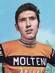 Merckx holding a bicycle. His shirt says "Molteni Arcore", and his hair is slicked back.