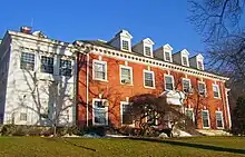 A wide brick building with dormer windows projecting from its roof and a white wooden wing on the left, seen from slightly downhill