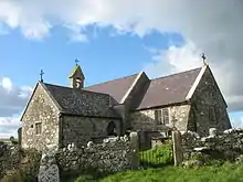 A stone church with slate roofs seen from the southeast. To the right is the chancel, to the left is a large transept, beyond which can be seen a bellcote