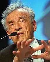 A photo of an elderly man in a suit speaking into a microphone