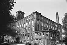 A five-storey cuboid factory composed of brick and windows. The view is from the ground at one corner of the factory. To the right, appearing from behind the factory, is a tall brick chimney. To the left are trees appearing black, and at their fringe, a square brick tower connected to the factory.