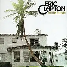 A house is shown along with a palm tree, on the right Eric Clapton holds his hands smiling, above him is the title and logo along with some trees