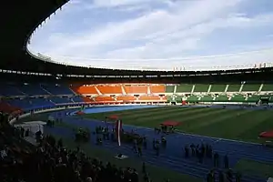 An oval shaped stadium with a blue athletics track and stands with red, blue, orange and green seating sections. One of the lateral stands is filled and more people are in the track. The sky is blue with clouds.