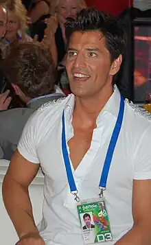 Seated, smiling young man in white wearing a backstage pass