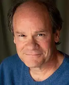 A white man with greying brown hair and a blue sweater is smiling into the camera.