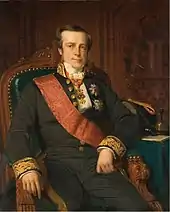 Portrait of a clean-shaven, square-faced man sitting in a chair. He wears a black suit with embroidered cuffs and a red sash over his right shoulder. Several awards are pinned to his coat.