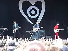 All three brothers are on stage playing guitars. Audience members have hands raised. Keyboards, speakers and other stage equipment are behind the men.