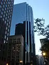 Ground-level view of a 40-story skyscraper with an all-glass facade and several setbacks. In the facade is a reflection of a dark brown building with lighter windows, and near the bottom of the skyscraper is a small white building with dark windows