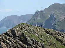 Ricky peaks and cliffs topped by greenery, with a small section of blue see visible to the left.