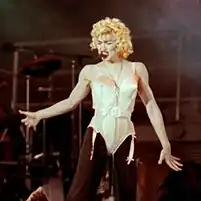 The image of a young blond woman. She is wearing a beige corset and black pants. She has blonde curly hair and has a headset microphone to her mouth.