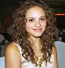 A smiling, dark-eyed, olive-skinned young woman with wavy dark brown hair that goes down below the shoulders, wearing a sleeveless light beige top and a gold necklace