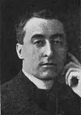 Black and white photograph of a man wearing a Roman collar from the mid-chest up. He faces the camera and touches his index finger to his temple.