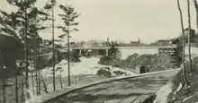 "Black and white photo of a road meandering alongside a river rapids, eventually crossing the top of the rapids along a steel arch bridge."