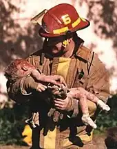 A firefighter is holding a dying toddler in his arms, and he is looking down at her. The toddler has blood on her head, arms, and legs, and is wearing white socks.