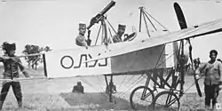 Two men seated in a World War I-era biplane, surrounded by technical personnel.