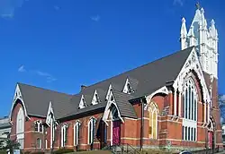  A brick church, seen from a corner and looking uphill, with a tall white steeple. One of its front windows has been boarded up.